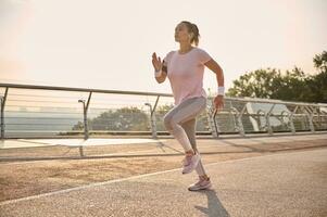 Determined strong athletic middle aged Hispanic sportswoman in sportswear during her morning run, running on a treadmill along a city bridge with beautiful nature in the background photo