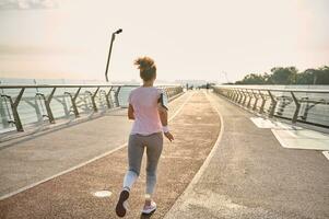 espalda ver de joven atlético mujer, persona que practica jogging, corredor practicando deporte al aire libre, corriendo rápido a lo largo el puente terminado el amanecer cielo y río antecedentes. sano estilo de vida concepto rutina de ejercicio trotar actividad foto