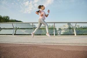 Active and healthy lifestyle, outdoor cardio, weight loss, running concept. Side full length of a determined active athletic woman exercising outdoor, running fast along the city bridge treadmill. photo