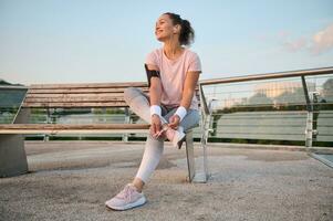 contento Encantado caucásico hembra atleta deportista en rosado camiseta con teléfono inteligente poseedor y terry pulseras descansa después Mañana trotar a amanecer sentado en un de madera banco en un urbano ambiente foto