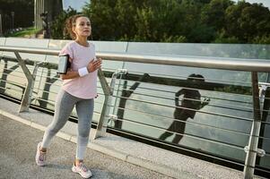 Determined strong athletic middle aged African American woman jogger in sportswear during her morning run, running on a treadmill along a city bridge with beautiful nature in the background photo