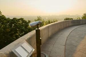 Tourist coin telescope. Metal stationary binoculars standing on a high hill on the viewpoint, for observing the nature of a beautiful old historic city overlooking the river photo