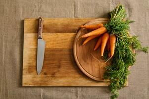 Flat lay of a big bunch of fresh carrots with green tops and a kitchen knife on a wooden cutting board on a linen tablecloth. Food background of seasonal raw vegetables photo