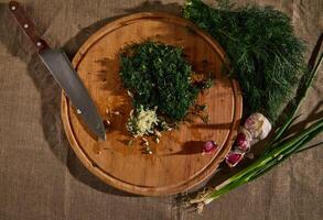 Overhead view of a bunch of fresh moist dill on a linen tablecloth and garlic next to a wooden cutting board with a kitchen knife and chopped garlic and dill. Seasonal greens food background. Flat lay photo