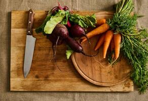 Flat lay. Seasonal raw vegetables. Bunches of fresh organic beets, baby carrots with tops and kitchen knife on a wooden cutting board. Vegan food background photo