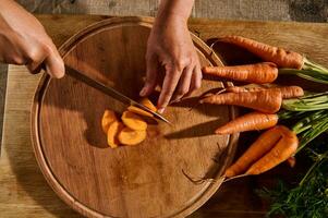 Overhead view of hands chopping, slicing fresh raw baby carrot on wooden board. Close-up of hand with knife cutting vegetable. photo