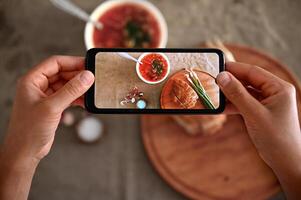 Top view of hands holding smartphone and photographing a bowl with freshly cooked Ukrainian traditional soup- Borscht with fresh green raw onion . Mobile hone in live view mode photo