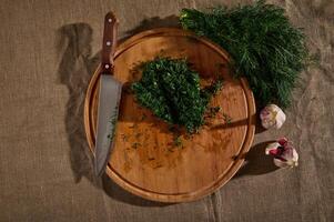 Top view of a bunch of fresh moist dill on a linen tablecloth and garlic next to a wooden cutting board with a kitchen knife and chopped dill. Seasonal greens food background. Flat lay. photo