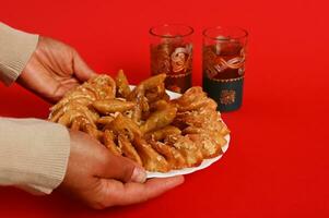 de cerca de mujer manos servicio un plato de delicioso oriental tradicional marroquí dulce postre y menta té en un hermosa Arábica estilo vaso. aislado en rojo antecedentes. foto