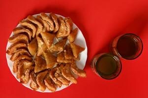 Flat composition with oriental moroccan dessert on plate next to two glasses in arabic style on red background with copy space. Arabic traditional oriental sweets on the festive table photo