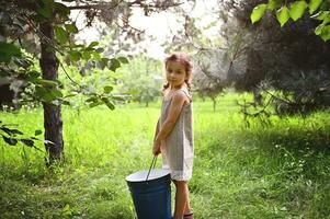 Beautiful little girl in a linen dress stands in the garden with a large blue metal bucket in her hands photo