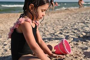 Preschooler girl playing with rake and pink toy bucket with sand, building sandy shapes and castles, enjoying sunbathing and summer vacations. Happy childhood, wellness and recreation concepts photo