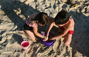 Children enjoying construction of sand figures on the beach. Boy and a girl fill a toy plastic bucket with wet sand to build sand castles. Brother and sister having fun during summer vacation photo
