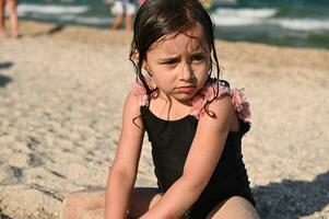 disparo a la cabeza retrato de linda trastornado bebé niña con mojado pelo y traje de baño, sentado en el arenoso playa en el mar antecedentes. niños emociones verano fiesta concepto foto