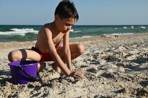 Handsome teenager in red swimming trunks playing on the sandy beach. Adorable child builds sand figures with wet sand against the background of the sea. Summer holidays concept photo