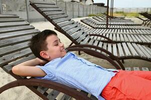 Happy teenage boy lying down on a wooden chaise lounge. Handsome child resting on the deck chair during summer country vacations. photo