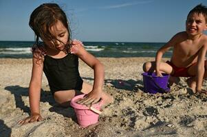 linda niña llena un rosado el plastico juguete Cubeta con arena a crear estatuillas y construye un arena castillo mientras jugando junto a su hermano en contra el fondo de el mar. foto
