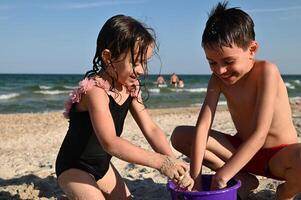 Brother and sister, friends,boy and girl filling plastic toy bucket with wet sand and building sandy figures. Happy children playing on the beach at sea background, enjoying summer vacations photo