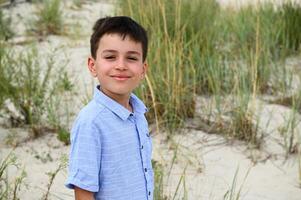 Close-up of the face of a cute smiling teenage boy standing against the backdrop of steppe thickets and sandy soil. Handsome child cute smiles looking at the camera. Headshot of adorable kid photo