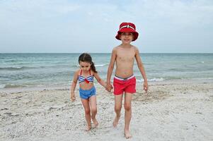 Beautiful kids walk along the beach during summer holidays. A handsome boy in a red summer hat and swimming trunks leads his sister by the hand against the backdrop of a seascape. photo