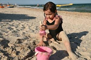 Active summer holidays concept. Baby girl playing with plastic toy bucket and shovel sitting on the sand, having sunbathing on the sea background photo