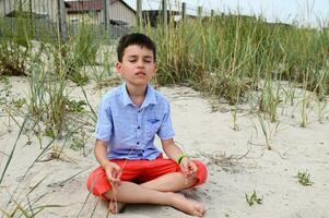 A handsome schoolboy in orange shorts sits on the sandy ground in a lotus position and meditates calmly against the backdrop of nature. Relaxation, serenity and wellbeing concept. photo
