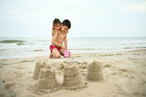 hermoso chico llena Cubeta con mar arena mientras su mas joven hermana suavemente abrazos a él. bonito niña abrazos su hermano linda mientras jugando playa juegos. borroso construido castillos de arena en el primer plano foto