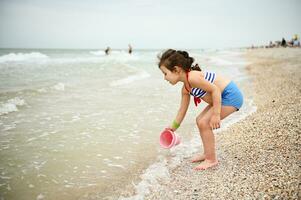 Pretty baby girl in a swimsuit collecting sea water in a pink toy bucket. Cute child playing on the beach. Healthy rest, recovery and recreation of children during the summer holidays. photo