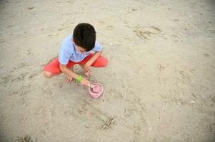 parte superior ver de un feliz, hermoso y adorable chico, niño, niñito relleno juguete Cubeta con arena para edificio castillos de arena a el playa. naturaleza, marina antecedentes. verano ocio actividades. foto