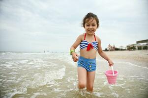 Pretty baby girl with a pink toy bucket in her hand , walking along the seacoast, enjoying sunbathing, outdoors recreation and cute smiling looking at the camera . photo