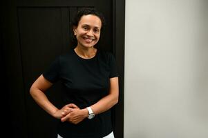 Middle aged mixed race woman, physiotherapist at a spa wellness center, wearing a black t-shirt, smiling cutely at the camera, standing in front of the door of her massage parlor photo