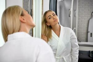A gorgeous woman in a white robe touches her blonde hair, admiring her reflection in the mirror. Beauty portrait of attractive woman with perfect smooth skin photo