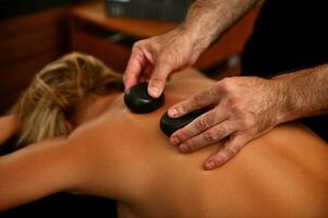 Masseur placing therapeutic hot stones on woman's back. Ayurvedic hot stone massage in modern spa salon. Body care concept photo