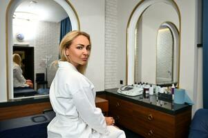 Young european woman in white robe looking over shoulder at camera while relaxing in beauty spa salon photo