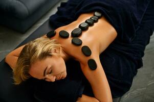 Beautiful European young blonde woman with beautiful face lying on a massage table with hot lava stones on her back along her spine receiving Ayurvedic treatment in a wellness spa clinic photo