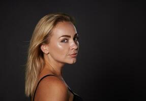 Close-up of confident blonde beautiful middle aged woman with healthy tanned clean glowing skin in black bra looking at camera standing three quarters on dark background with copy space. Studio shot photo