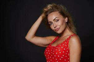 Attractive provocative sexy blonde woman in red swimsuit holding her hand on head and touching her flying healthy shiny hair, looking at camera isolated over black background with copy ad space photo