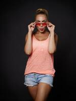 Beautiful blonde middle aged Caucasian woman looking through her pink sunglasses and showing tongue to the camera, posing over black background with copy space photo