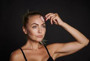 Face portrait of a pretty woman in black bra doing lymphatic drainage facial massage with a roller massager, isolated over dark background. Beauty spa treatment, copy space photo