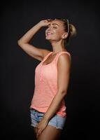 Attractive sexy young blonde woman with tanned skin, aesthetic fit body in denim shorts, pink top and sunglasses posing over black background with copy ad space photo