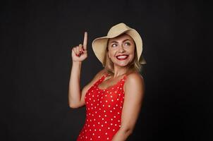 Happy cheerful stunning blonde woman in red outfit with white polka dots and straw summer hat smiles beautiful white toothy smile, pointing up finger at copy space on black background photo