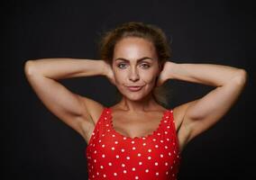 Beautiful blonde middle aged Caucasian woman in red swimsuit with white polka dots confidently looking at camera posing holding hands behind her head against black background with copy ad space photo