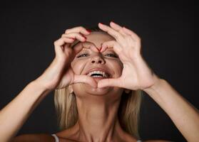 Close-up portrait of a cheerful good-looking blonde Caucasian woman smiles with beautiful healthy toothy smile looking at camera through a finger frame in heart shape, isolated over black background photo