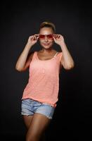 Beautiful sexy young blonde Caucasian woman with tanned skin, aesthetic fit body in denim shorts, pink top and sunglasses posing over black background with copy space for advertisement photo