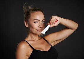 Beautiful Caucasian woman with tanned skin holding a tube of anti wrinkles smoothing anti puffiness under eyes cream, smiles looking at camera, isolated over black background with copy ad space photo