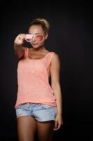 Attractive sexy young blonde woman with tanned skin, aesthetic fit body in denim shorts, pink top and sunglasses posing over black background with copy ad space photo