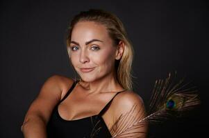 Close-up gorgeous smiling stunning middle aged blonde Caucasian woman with healthy fresh clean skin and wet blond hair holding a peacock feather, isolated over black background with copy ad space photo