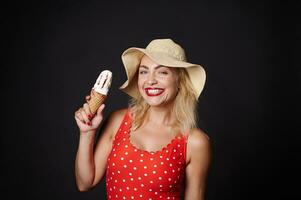 Beautiful woman in red swimsuit and summer straw hat with strawberry ice cream cone smiles with cheerful toothy smile against black background with copy ad space photo