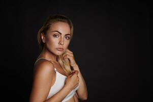 Confident portrait of an attractive young Caucasian woman with perfect clean skin and beautiful blond hair looking at camera standing against black wall background with copy space photo