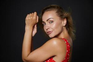 Portrait of a beautiful middle aged Caucasian woman with natural makeup and healthy glowing tanned skin, shiny flying blond hair, wearing red swimsuit isolated over black background with copy space photo